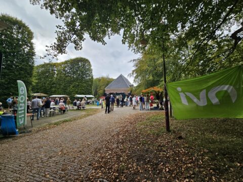 Buitenevenement met mensen, kraampjes, en bomen, groene spandoek en gebouw op de achtergrond.
