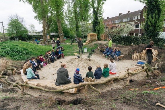 Kinderen zitten buiten in een cirkel op houten banken, omringd door groene bomen.