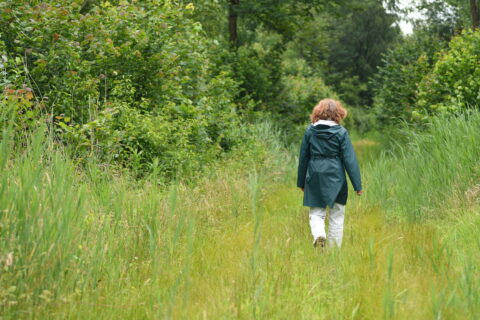 Persoon in regenkleding wandelt over groen pad omgeven door weelderige struiken en gras.