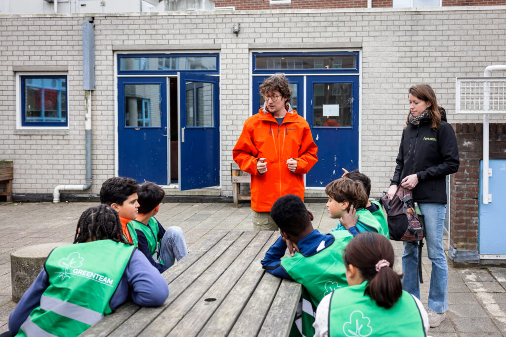 Man en vrouw begeleiden kinderen in groene hesjes, zittend aan een buitentafel bij een gebouw.