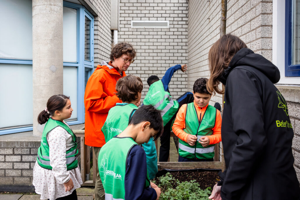 Kinderen in groene hesjes en een volwassene in een oranje jas verzorgen een klein tuintje bij een gebouw.