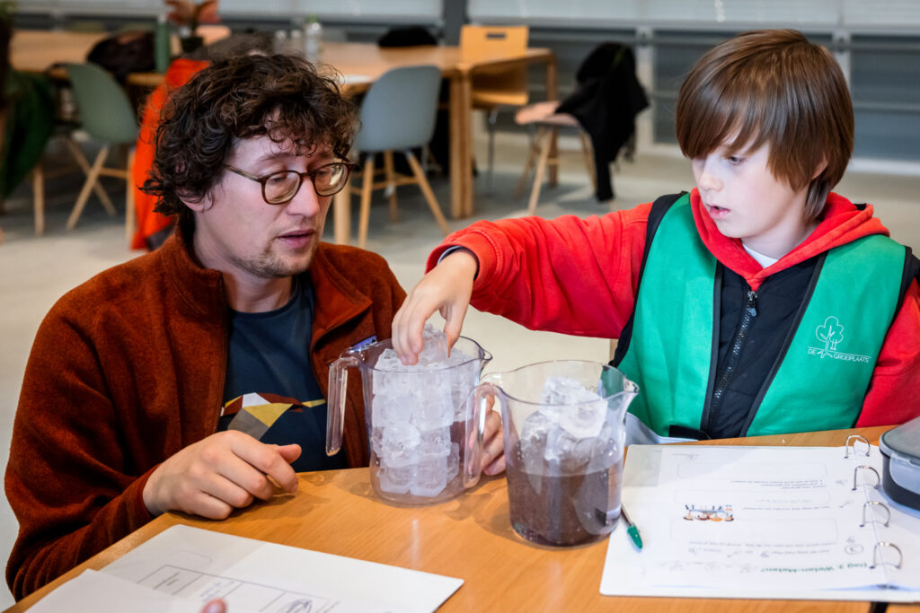 Volwassene en kind voeren een experiment met ijsblokjes uit aan een tafel met documenten.