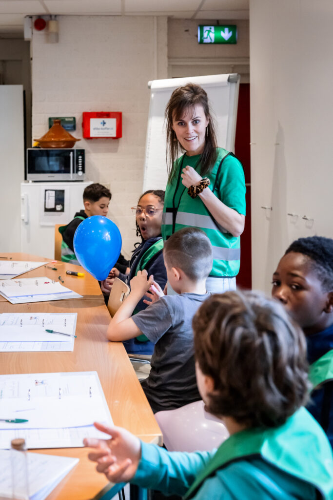 Vrouw begeleidt kinderen aan tafel met educatief materiaal en een blauwe ballon.
