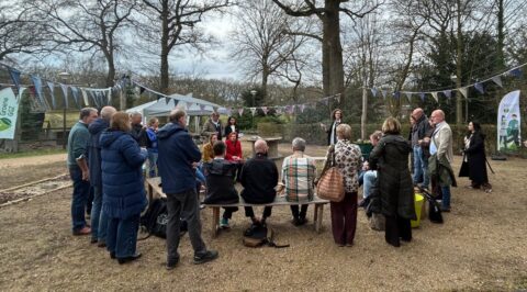 Groep mensen verzameld in een park met vlaggetjes en banners, omgeven door bomen.