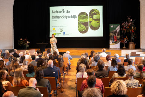 Vrouw presenteert over natuur in behandelpraktijk; mensen zitten in zaal, scherm met titel op achtergrond.