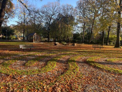 Park met herfstbladeren, bankjes, cirkelvormig pad en een houten gebouwtje op een zonnige dag.
