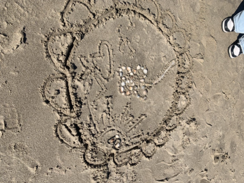 Zandtekening van een cirkel met schelpen en bloemmotief, naast voeten in slippers op het strand.