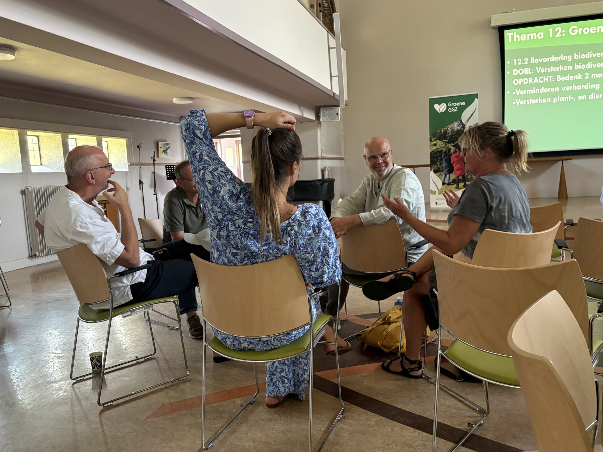 Een groep mensen zit in een kring en bespreekt thema's over biodiversiteit met een projectiescherm.