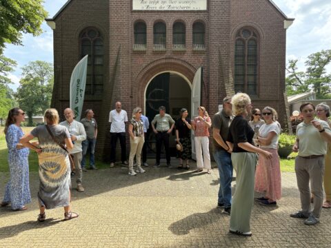 Groep mensen buiten een oud bakstenen gebouw op een zonnige dag, met open deuren en bomen rondom.