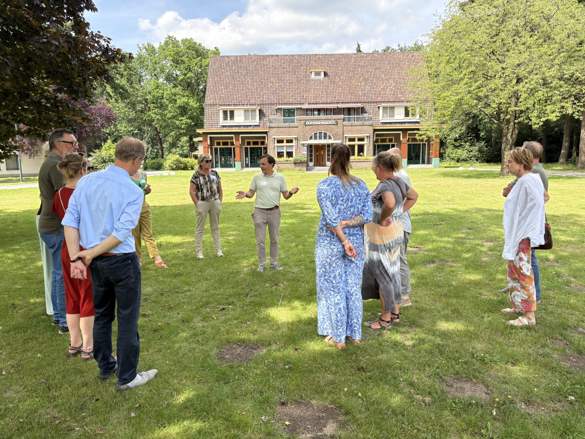 Een groep mensen staat in een kring op een grasveld voor een groot gebouw.