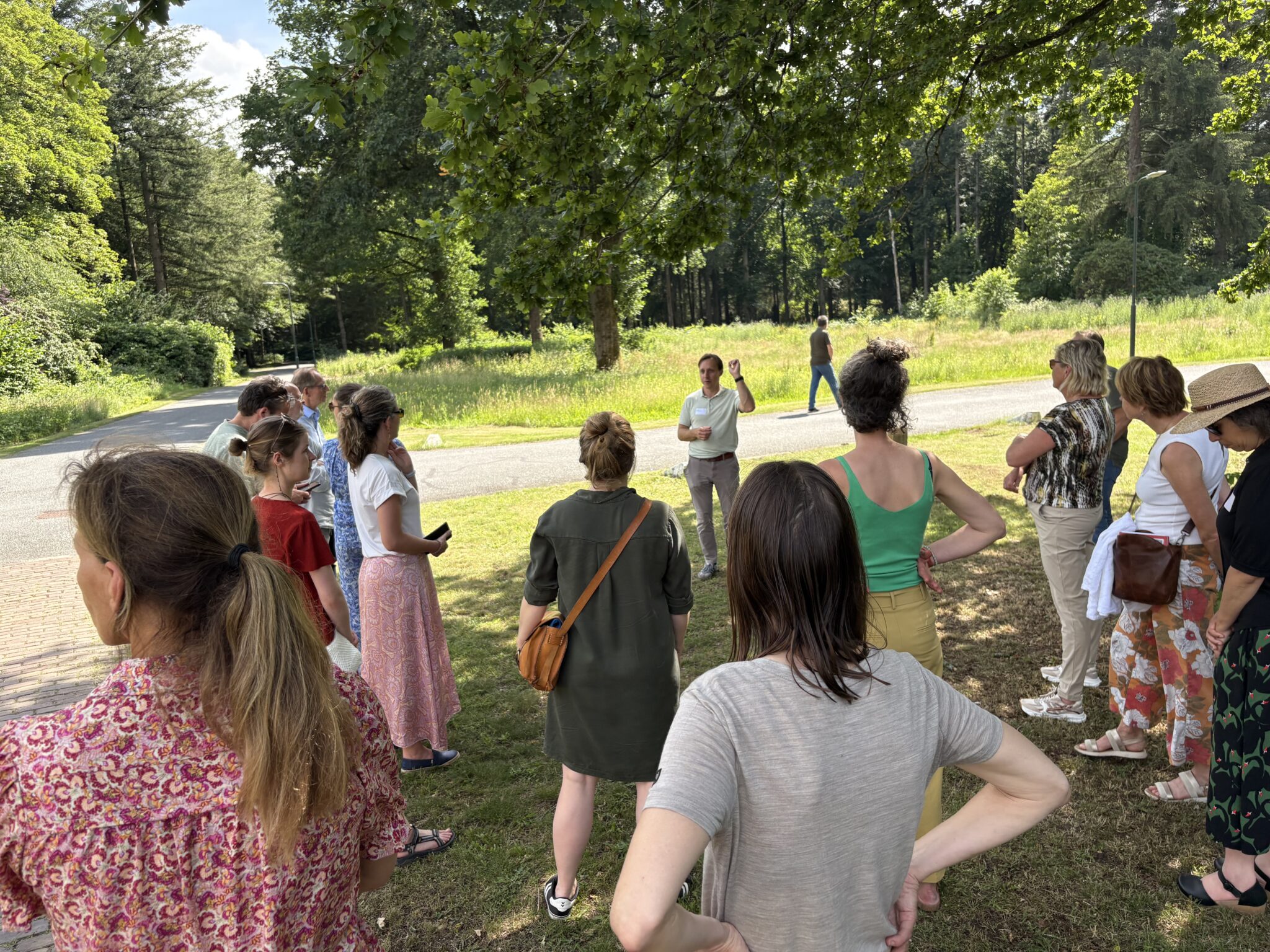 Groep mensen luistert naar een spreker in een bosrijk park op een zonnige dag.