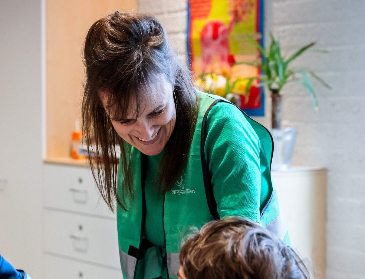 Vrouw glimlacht naar een kind in een klaslokaal, draagt een groen hesje.