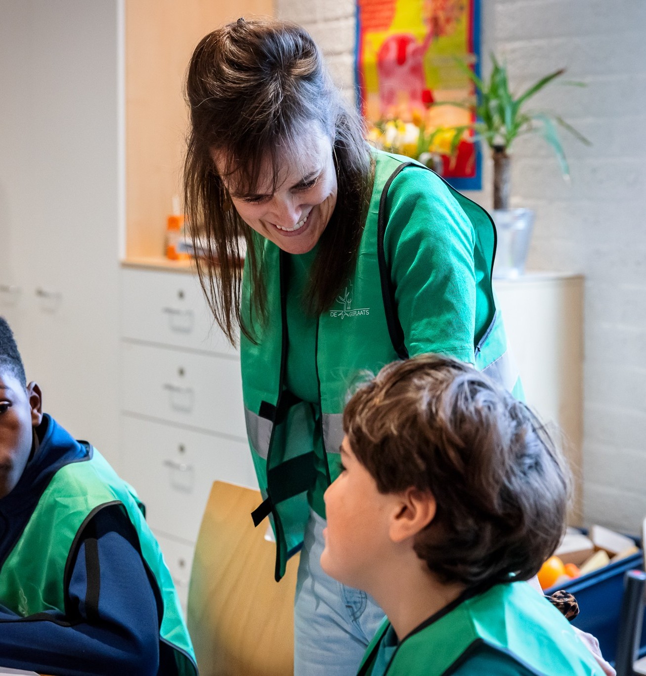 Een vrouw en twee kinderen in groene hesjes praten in een klaslokaal.
