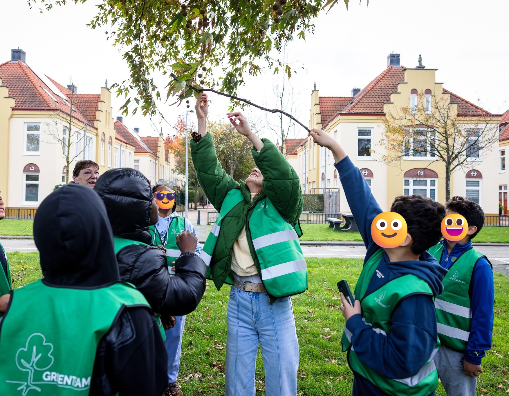 Groep kinderen in groene hesjes kijkt naar bladeren aan boomtak, voor huizen met rode daken.