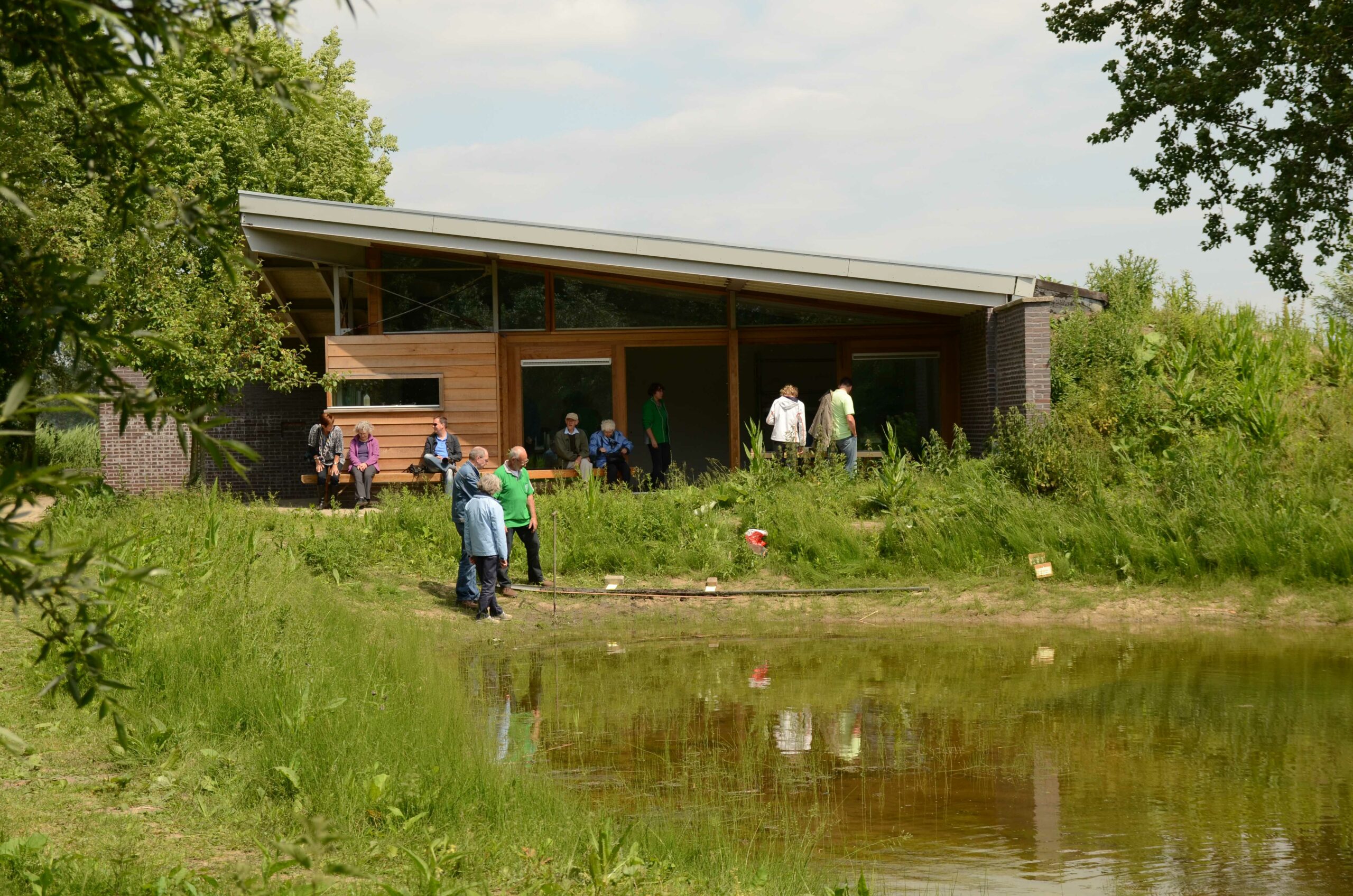 Mensen bij een modern houten gebouw aan een vijver, omgeven door groen en bomen.