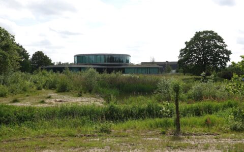 Rond gebouw met glazen gevels omgeven door groen en bomen onder een lichtbewolkte hemel.