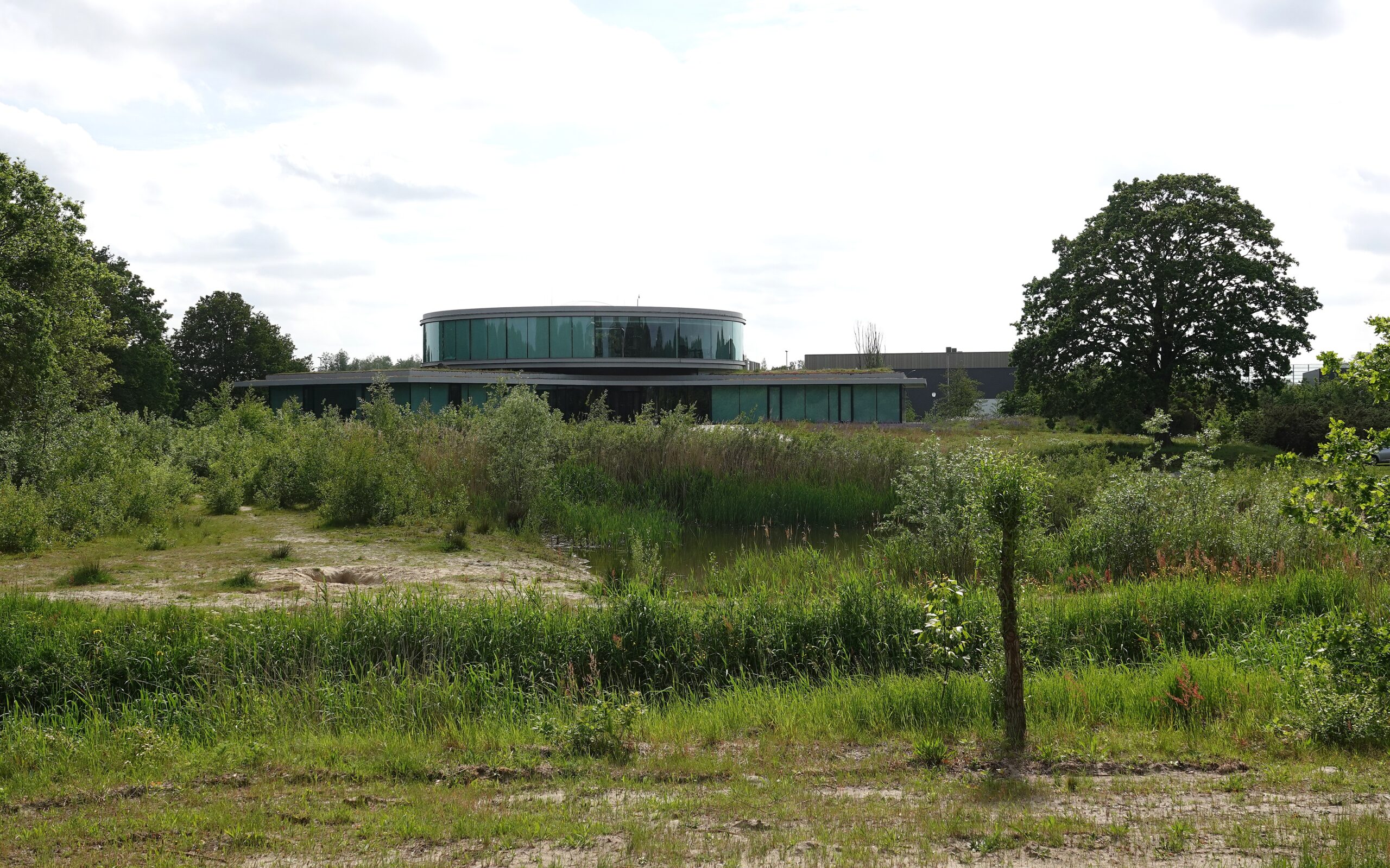 Rond gebouw met glazen gevels omgeven door groen en bomen onder een lichtbewolkte hemel.