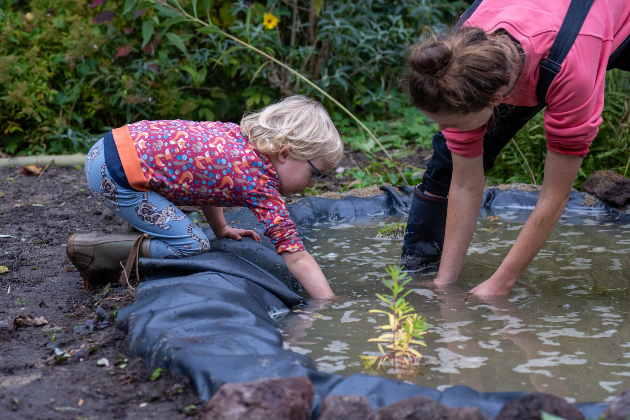 Een kind en volwassene graven in modder bij een vijver in een tuin.