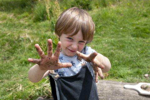 Een kind toont met plezier vuile handen buiten in een groene omgeving.