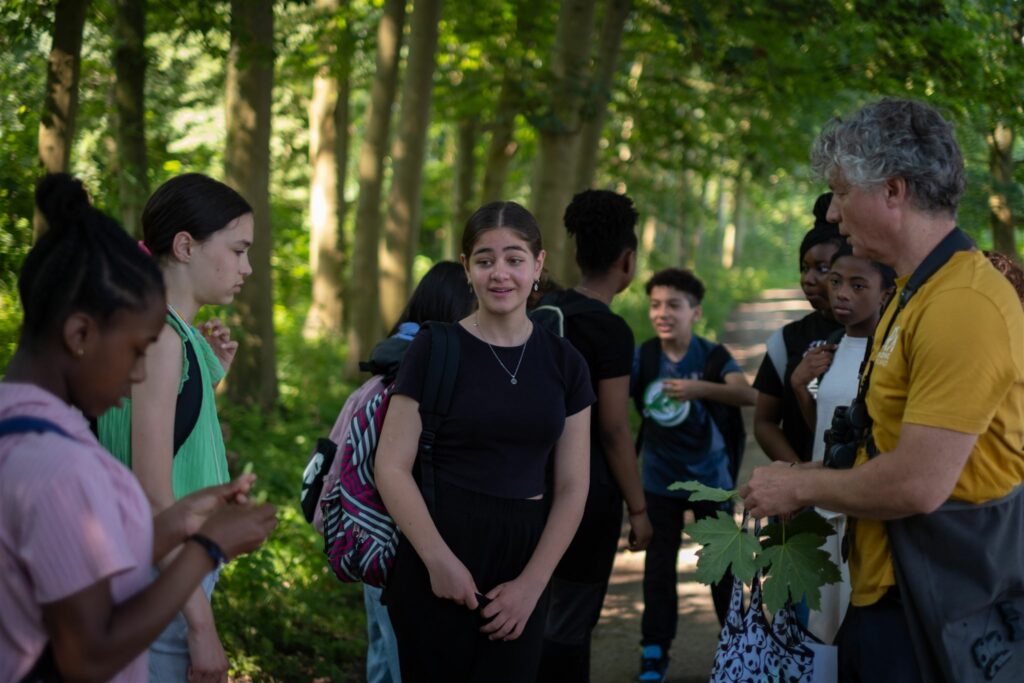 Groep jongeren met rugzakken in een bos, geleid door een man met bladeren in zijn hand.