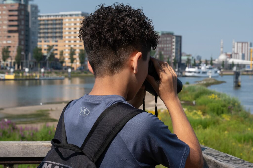 Jongen met verrekijker kijkt naar rivier met gebouwen op de achtergrond.
