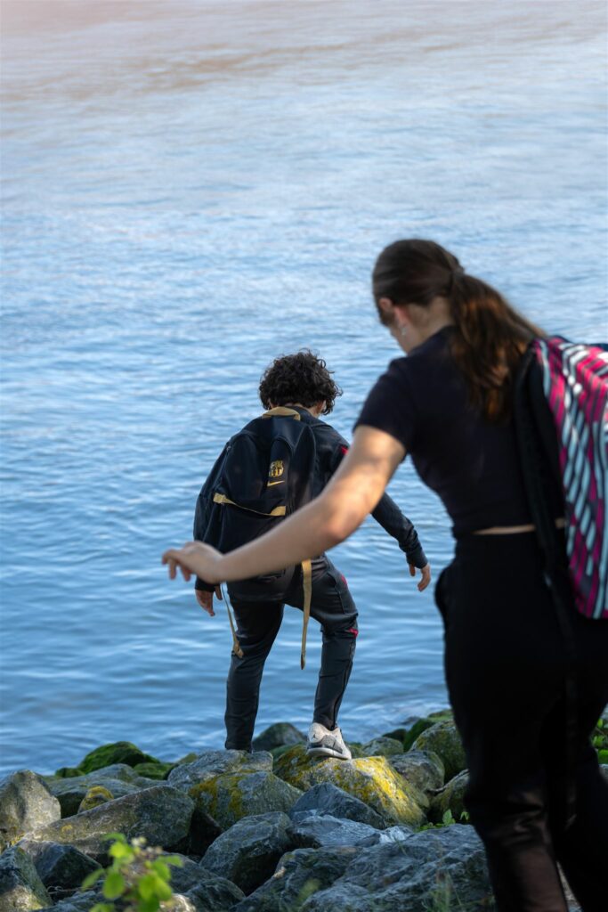 Twee mensen met rugzakken lopen over rotsen bij een waterkant.
