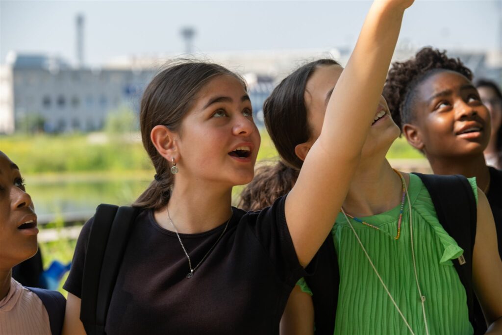 Jonge mensen kijken geïnteresseerd omhoog, buiten bij een gebouw, op een zonnige dag.