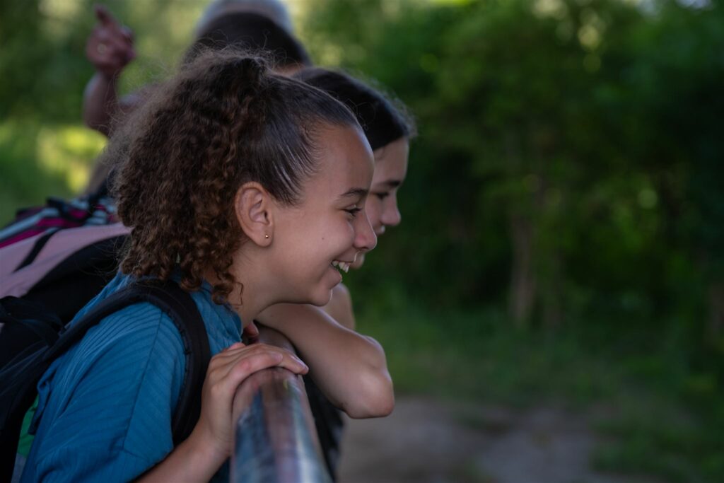 Meisjes glimlachen en leunen over een reling buiten, omgeven door groene bomen.