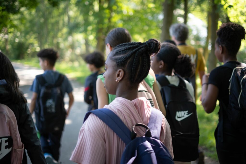 Een groep kinderen met rugzakken wandelt door een groen bos op een zonnige dag.