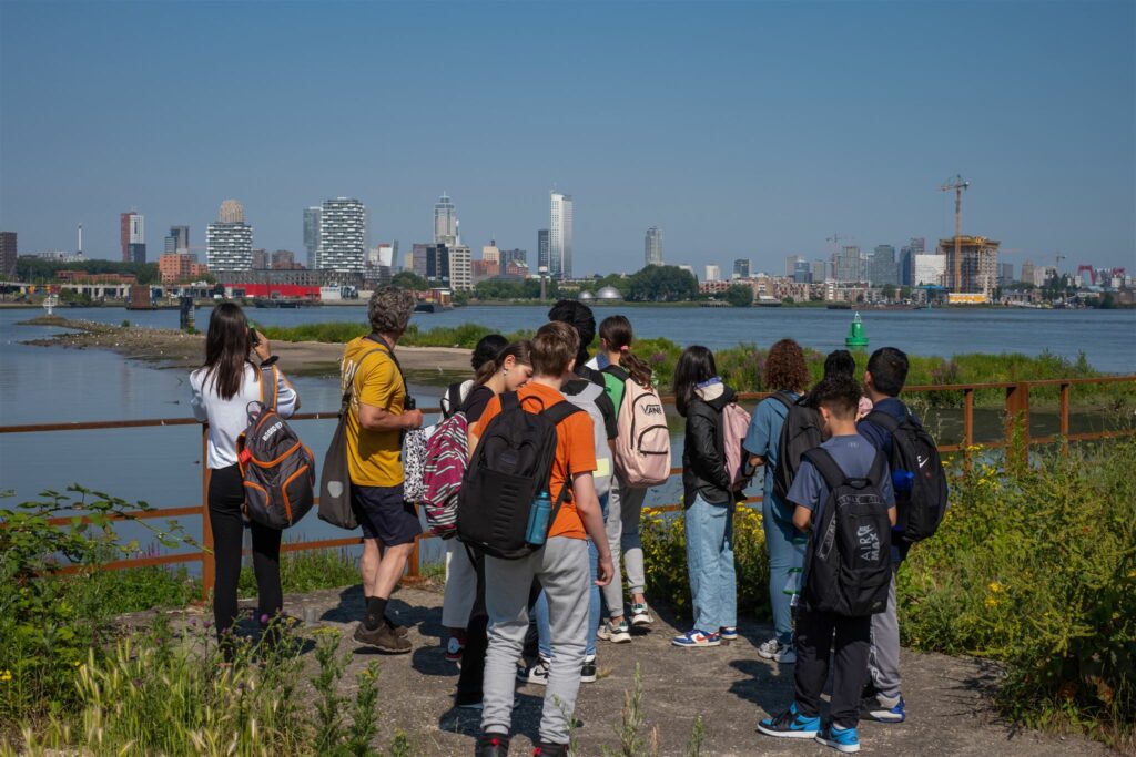 Groep mensen kijkt naar skyline over rivier, omgeven door groen, zonnige dag.