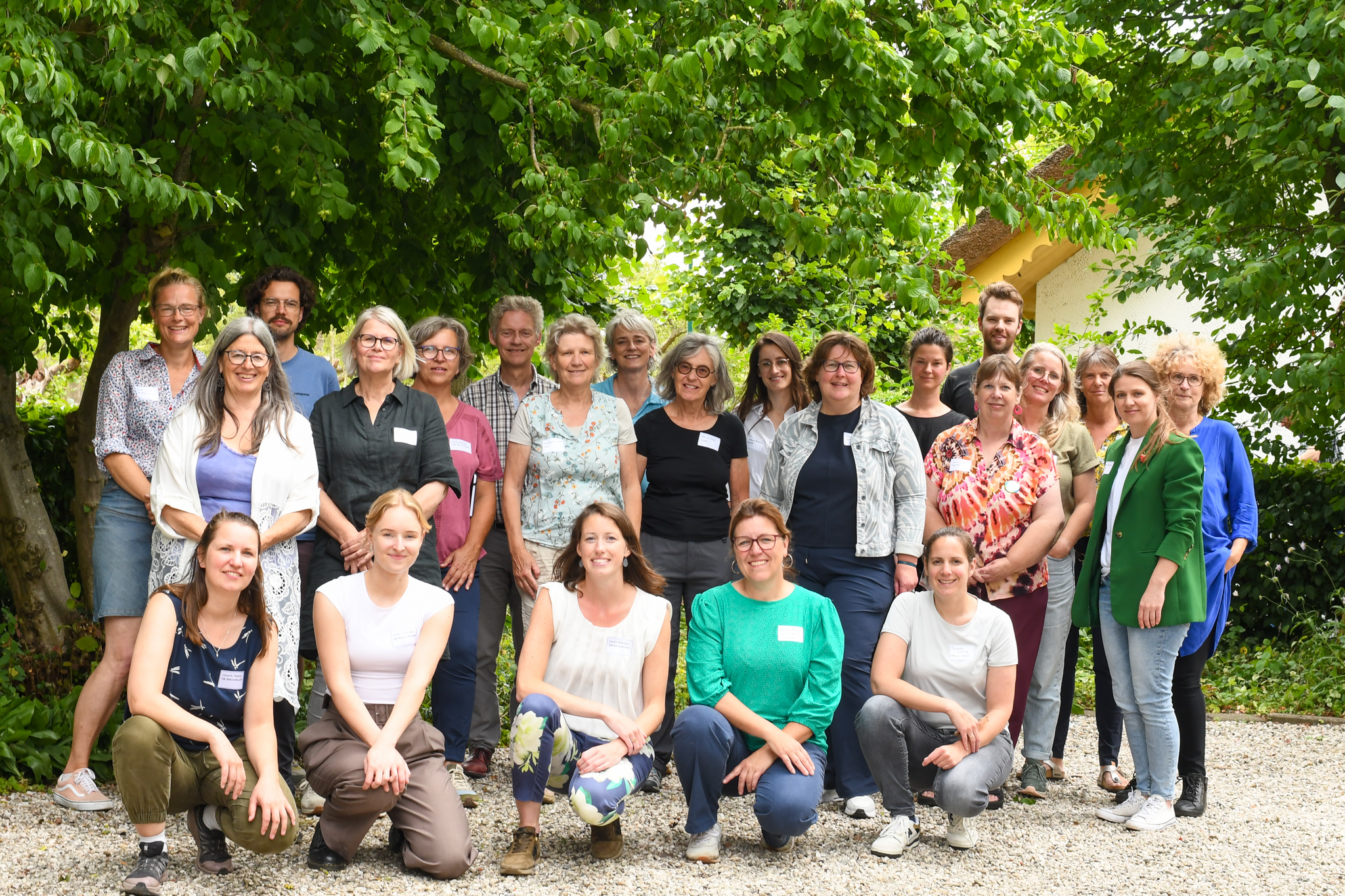 Groepsfoto van lachende mensen buiten onder groen bladerdak. Verschillende leeftijden en kledingstijlen.