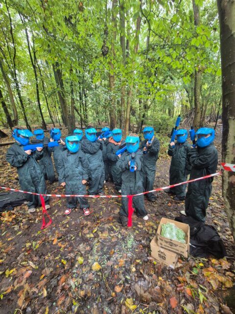 Groep mensen in blauwe paintballuitrusting in een bos, omringd door bomen en afzetlint.