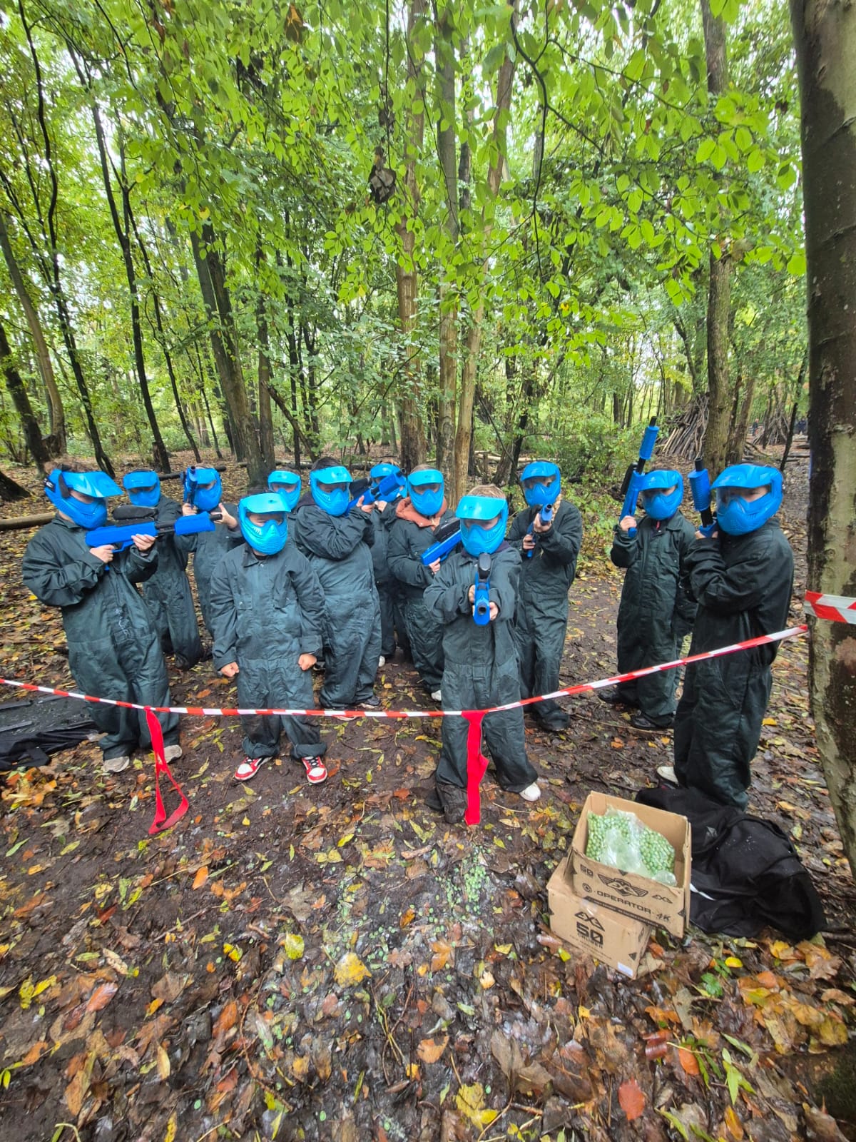 Groep mensen in blauwe paintballuitrusting in een bos, omringd door bomen en afzetlint.