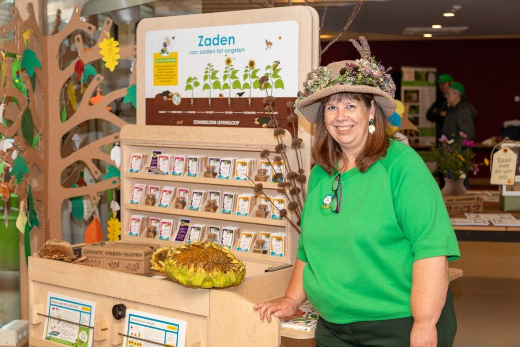 Vrouw in groene kleding bij zadenstand met zonnebloem en informatieborden over zaaien en oogsten.