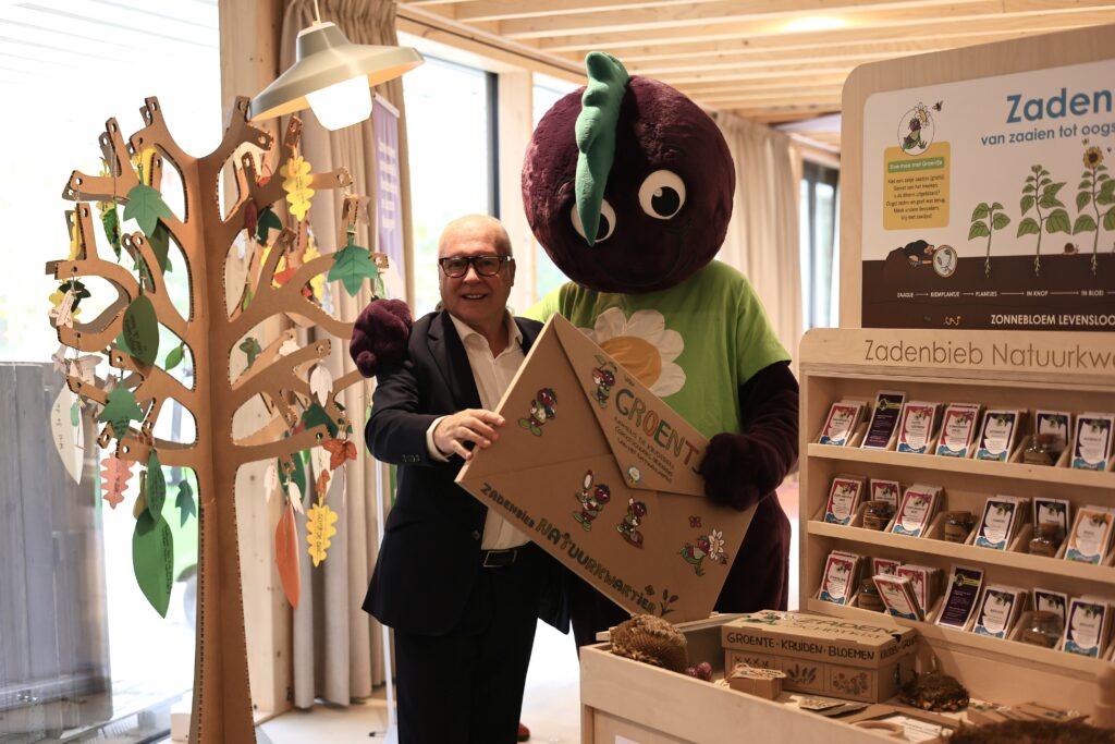 Man en mascotte met grote kartonnen envelop bij zaadbibliotheekstand met informatie over zaden.