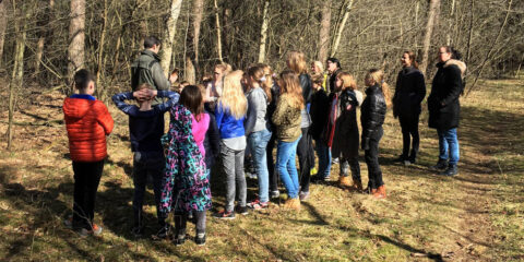 Groep kinderen en twee volwassenen in een bos tijdens een natuurwandeling onder leiding van een gids.