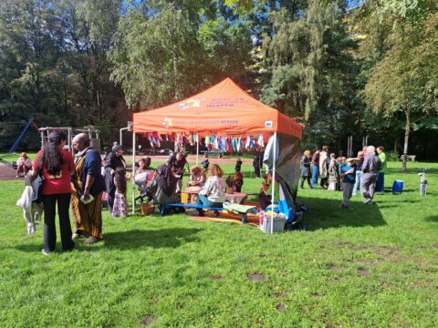 Bijeenkomst in park met oranje tent, mensen praten en kinderen spelen op zonnige dag.