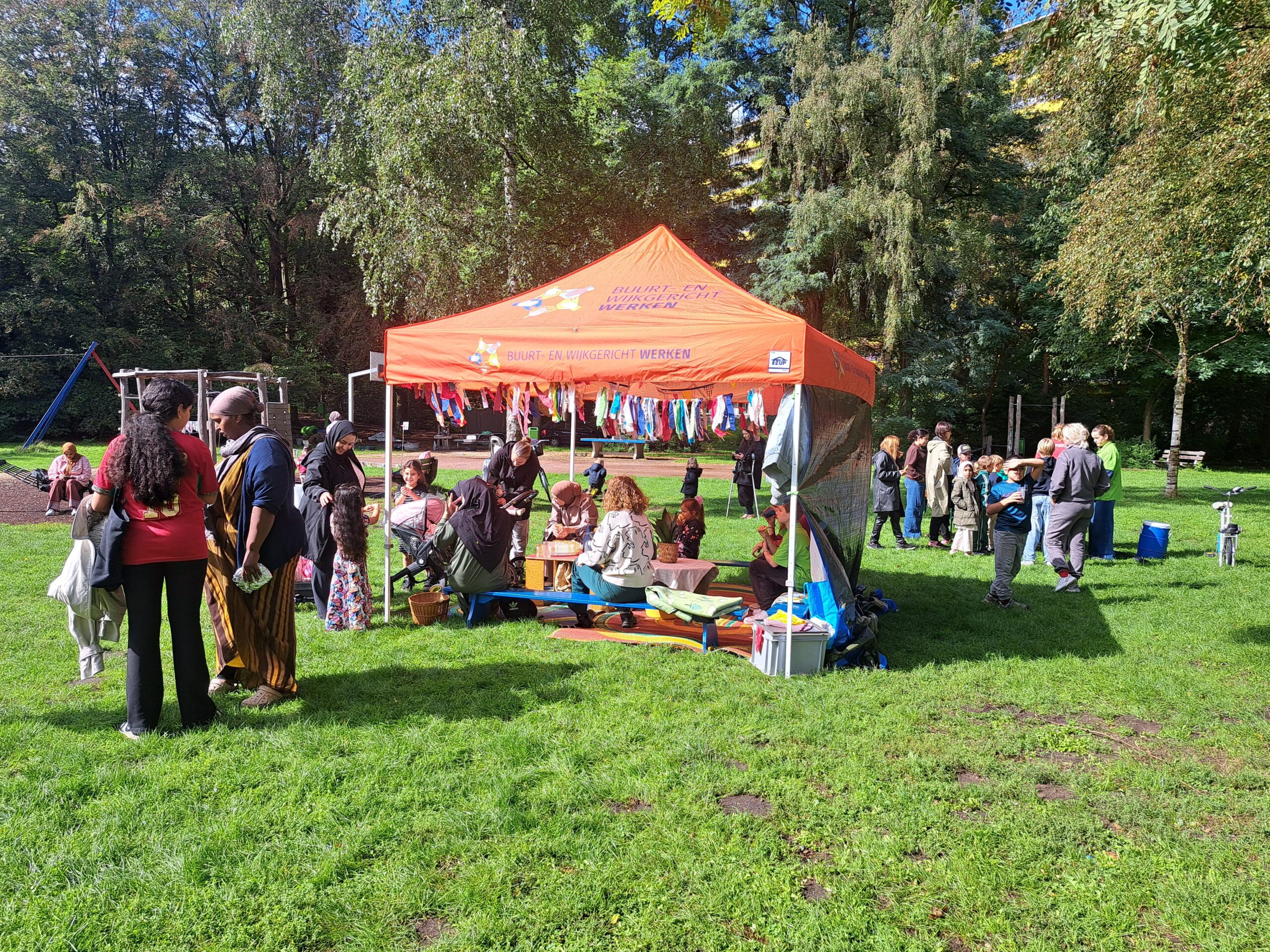 Bijeenkomst in park met oranje tent, mensen praten en kinderen spelen op zonnige dag.