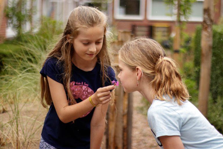 Twee meisjes in een tuin, één laat de ander aan een roze bloem ruiken.