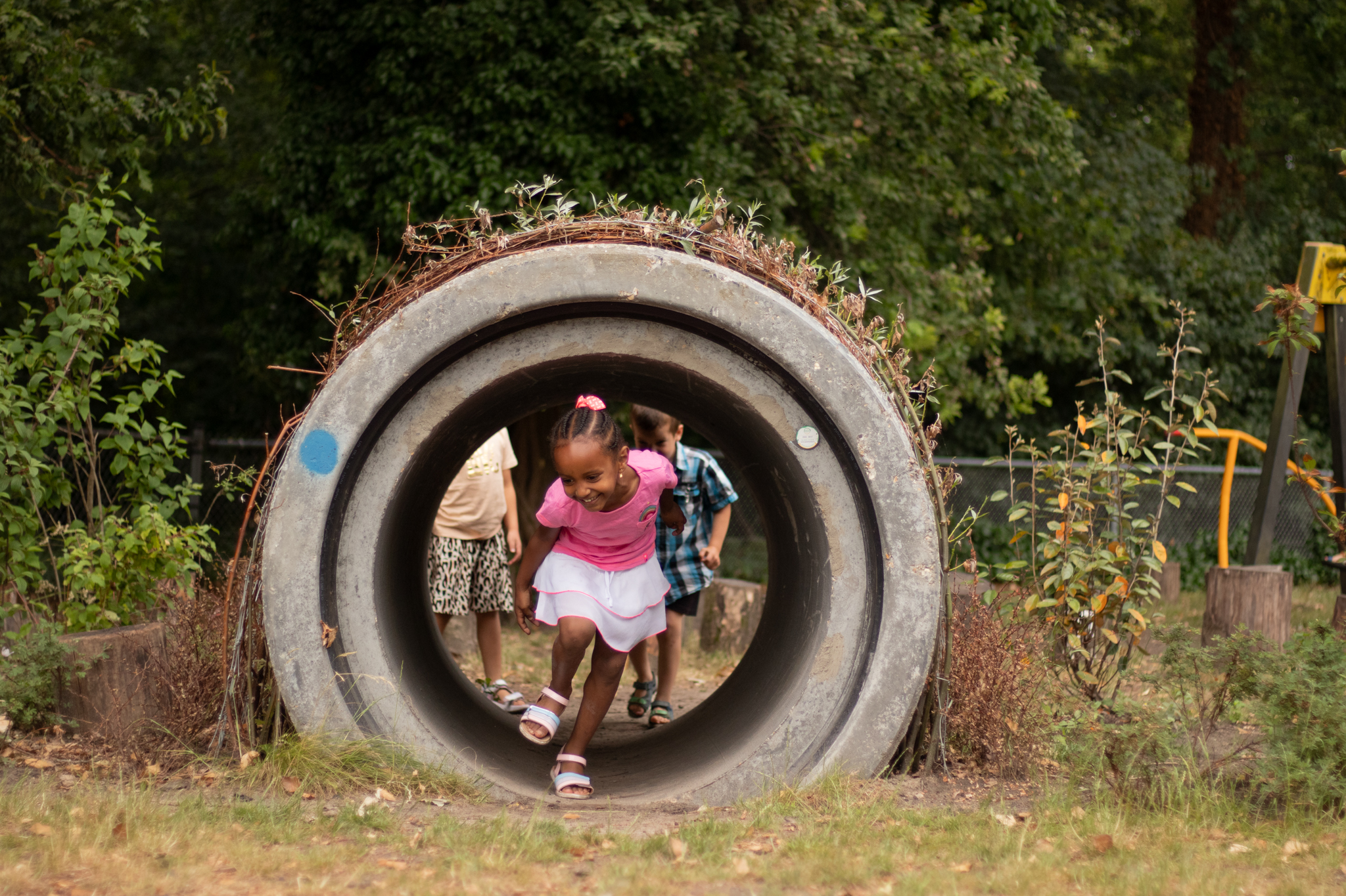 Kinderen rennen door een betonnen tunnel in een park met groene achtergrond.