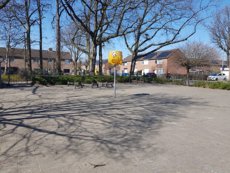 Plein met bomen, geel klimrek en woningen op de achtergrond onder een heldere blauwe lucht.