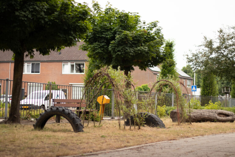 Speeltuin met autoband, bankje en boomstammen, omringd door woning en groen.