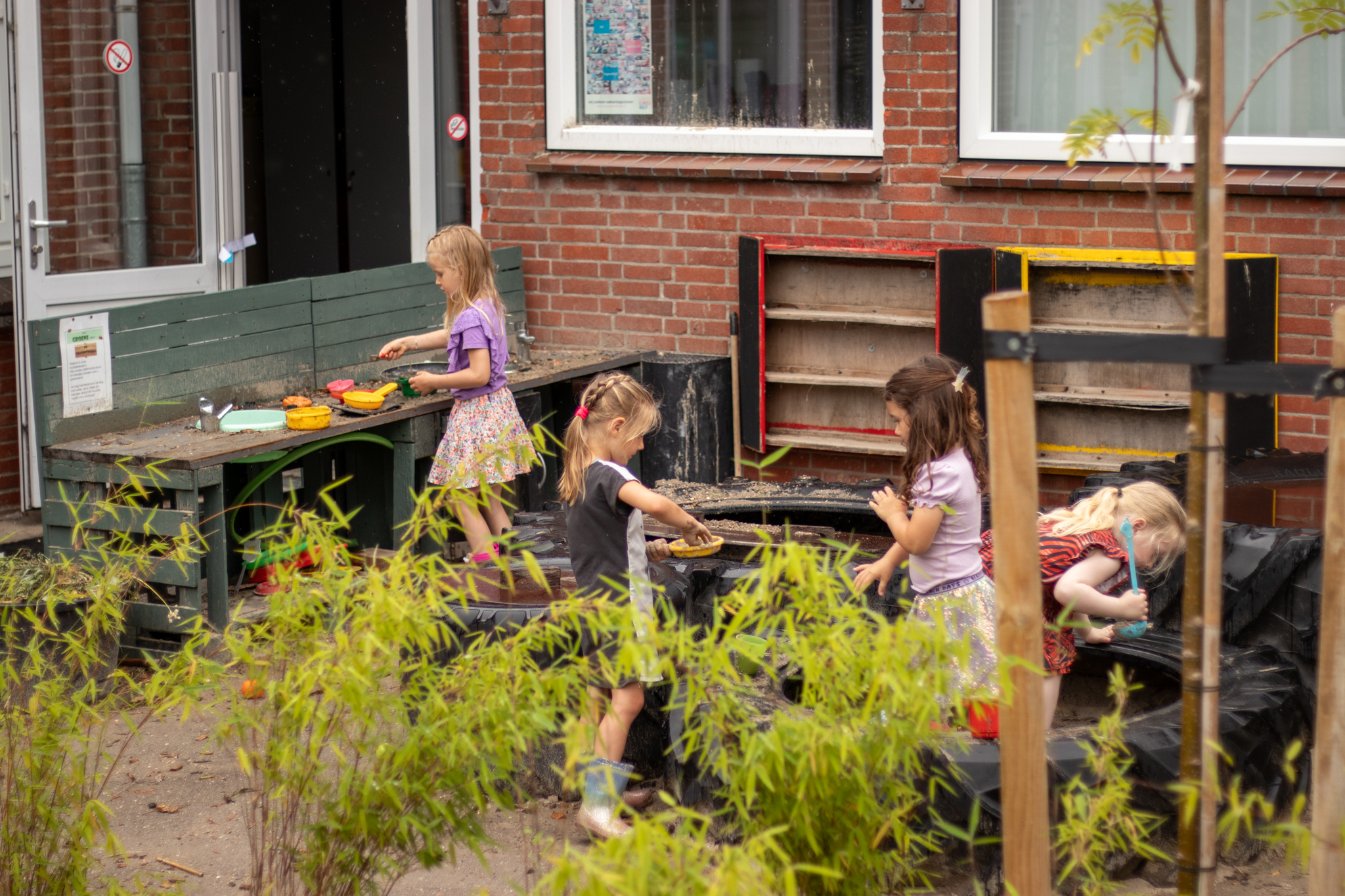 Kinderen spelen buiten bij een speelkeuken met bakjes en zand, omringd door groene planten.