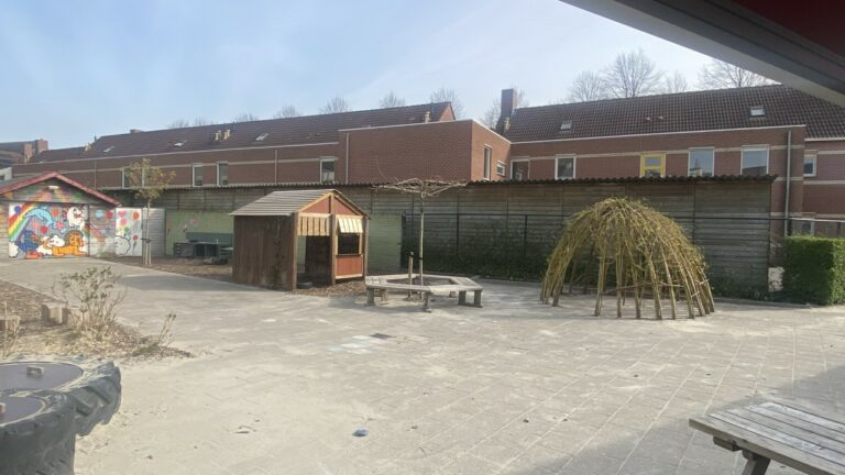 Speelplaats met houten hut, wilgenhut, kleurrijke muurschildering en bakstenen gebouwen.