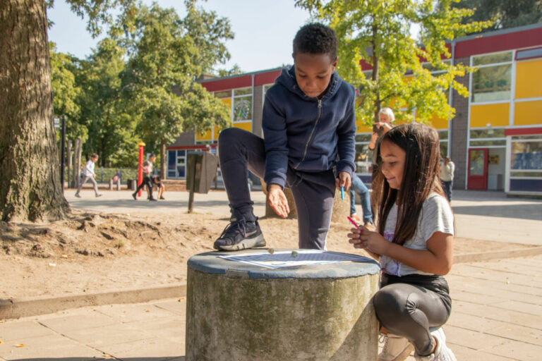 Twee kinderen spelen buiten bij een school op een stenen tafel, onder een boom.