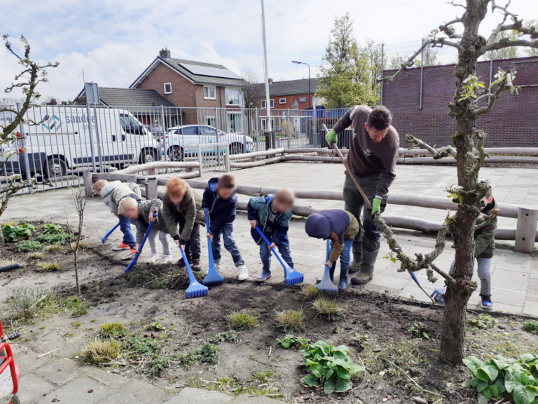 Kinderen en een volwassene harken samen in een tuin op een betegeld schoolplein.