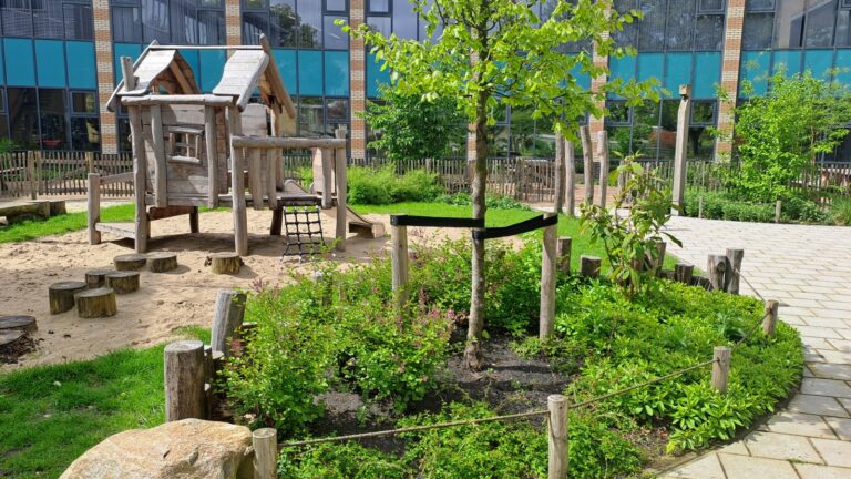 Speeltuin met houten speelhuis, zand, boom en groene struiken naast een gebouw met glasramen.