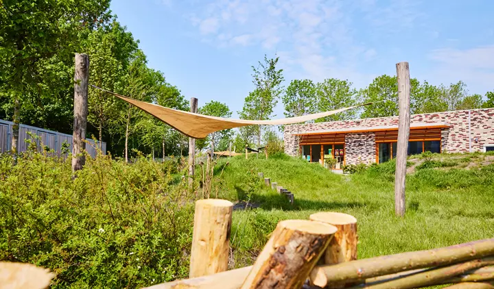 Een moderne stenen gebouw in groen landschap met een schaduwdoek en houten palen.