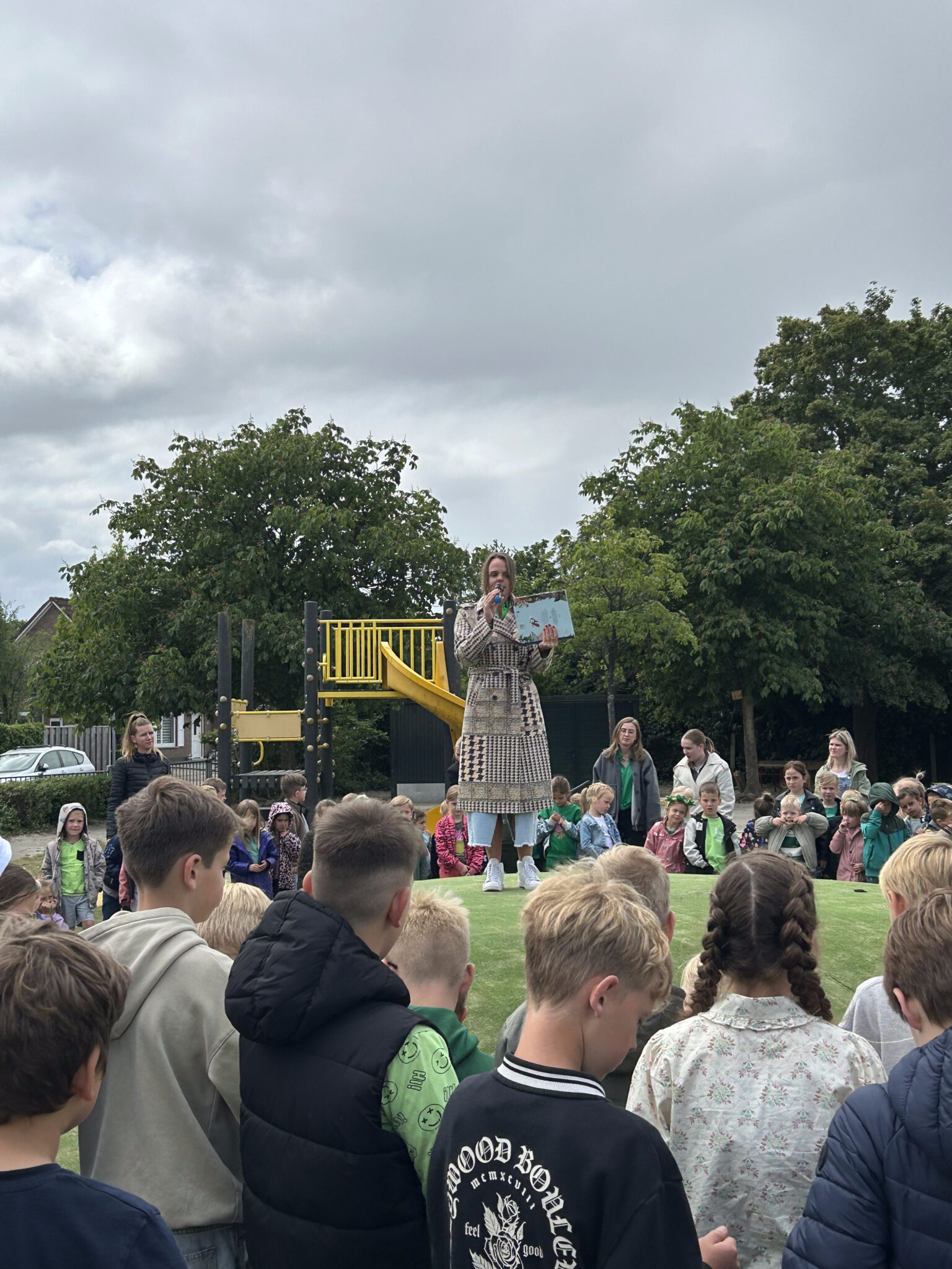 Vrouw spreekt voor een groep kinderen op een speelplaats met een glijbaan en bomen op de achtergrond.