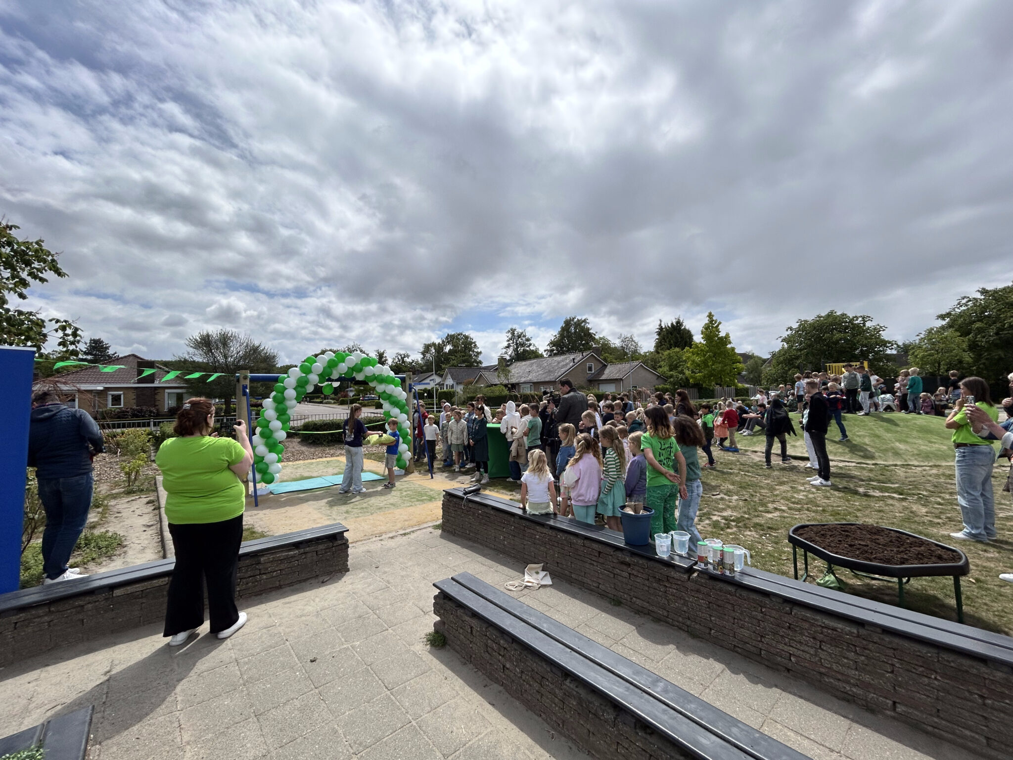 Buitenbijeenkomst met een menigte bij een boog van groene en witte ballonnen onder een bewolkte lucht.